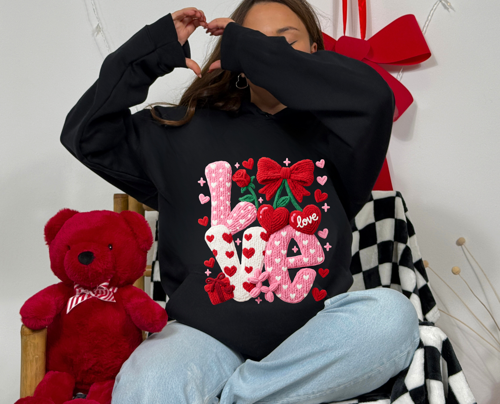 Person wearing a black sweatshirt with decorative text and graphics, sitting next to a red teddy bear.