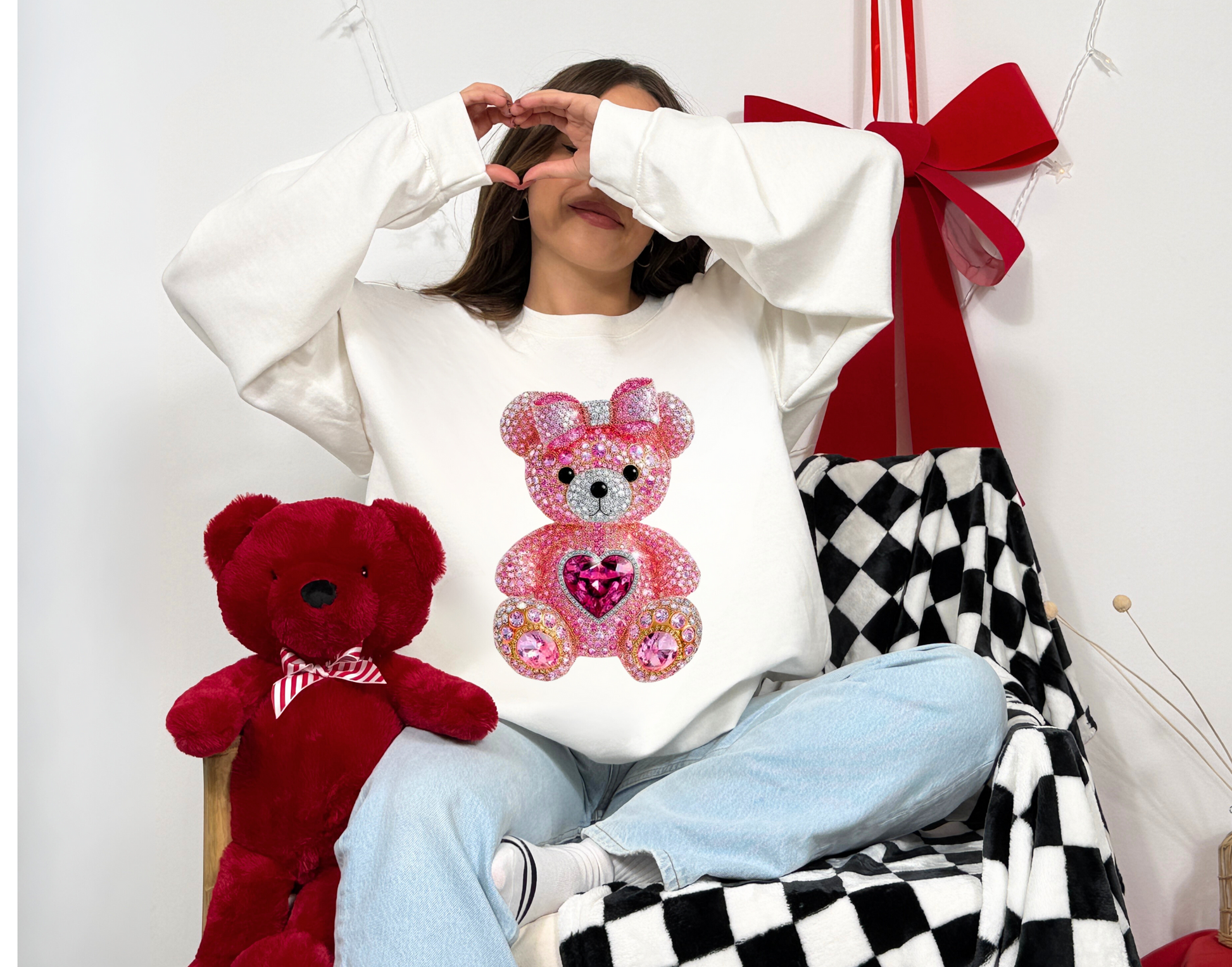 Person wearing a white sweatshirt with a pink teddy bear design, sitting on a checkered blanket with a red teddy bear and a red bow in the background.