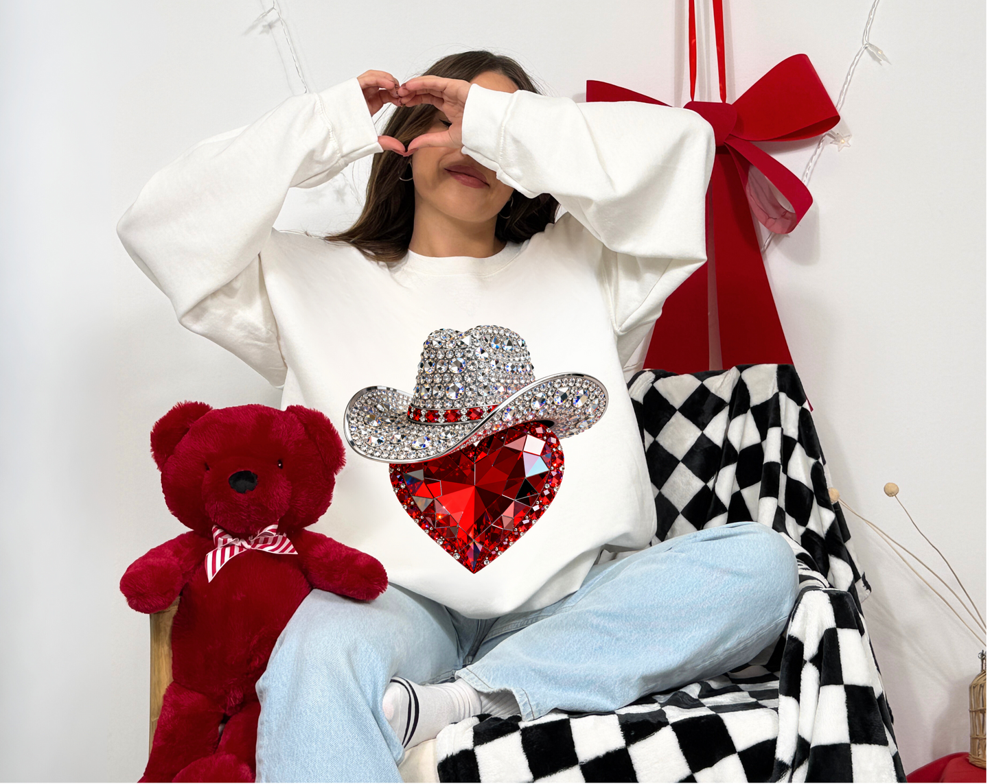 Person wearing a white sweatshirt with a decorative design, sitting next to a red teddy bear and a checkered blanket.