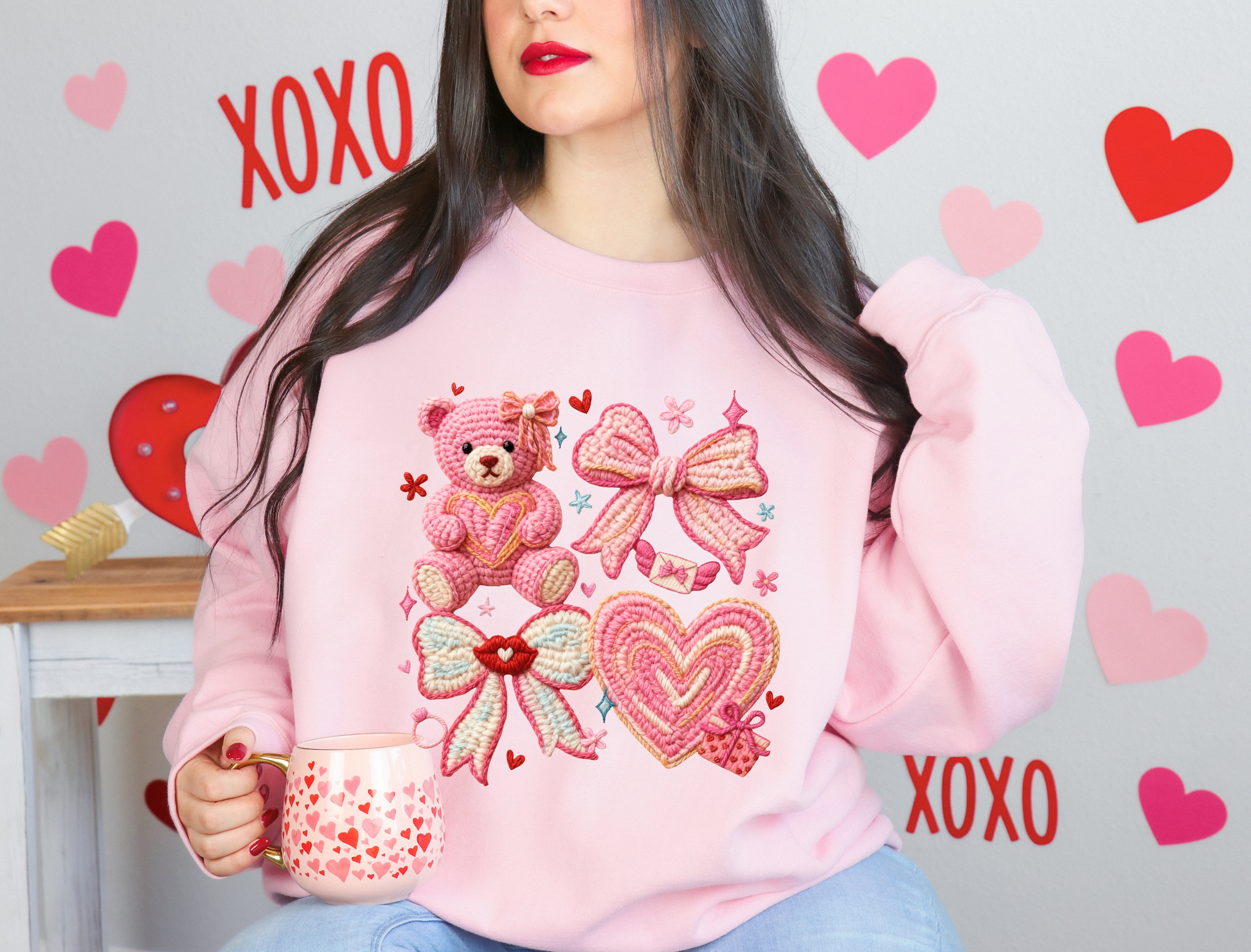 Person wearing a pink sweatshirt with teddy bear and bow designs, holding a mug, against a heart and 'XOXO' decorated wall.