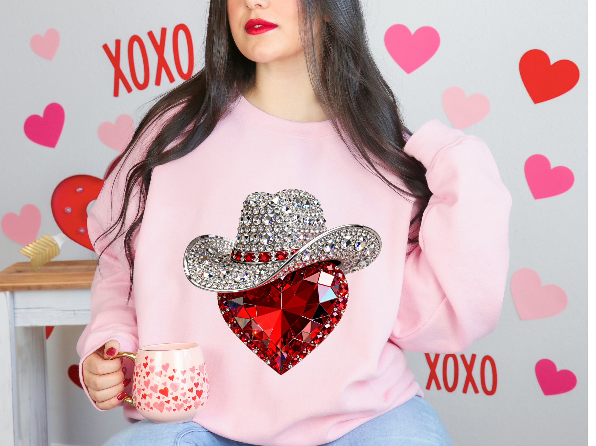 Person wearing a pink sweatshirt with a cowboy hat and heart design, holding a mug with hearts, against a heart-themed background.