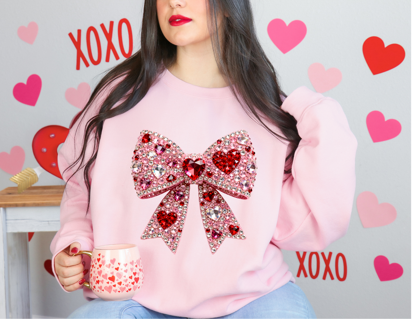 Person wearing a pink sweatshirt with a large bow design, holding a mug, surrounded by heart and 'XOXO' decorations.