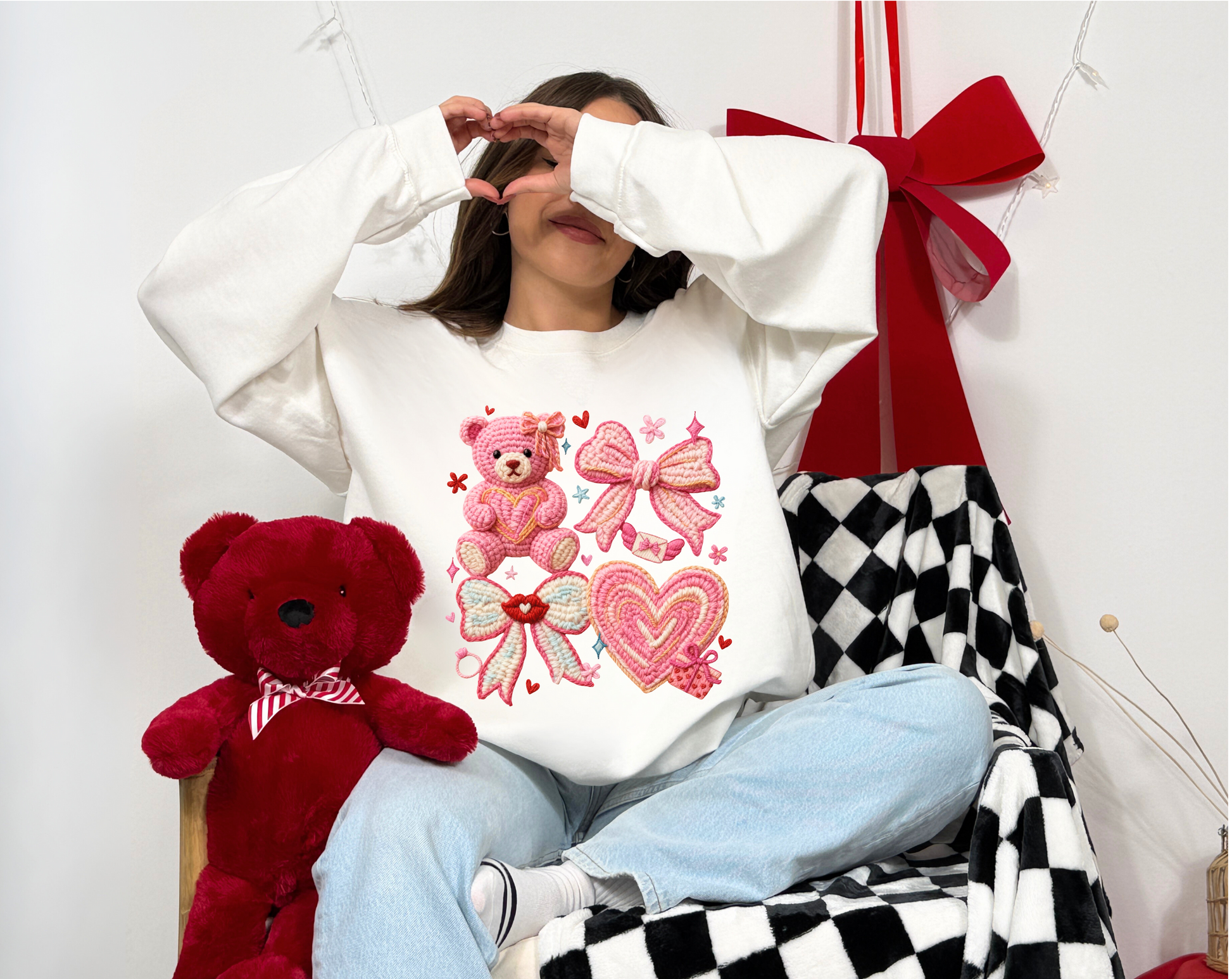 Person wearing a white sweatshirt with pink designs, sitting next to a red teddy bear on a checkered floor.